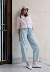 Woman leaning against wall wearing light pink cardigan, matching cap, light blue jeans, and white woven flats indoors.