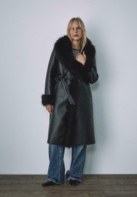 Czarny skórzany płaszcz z szerokim sztucznym futrzanym kołnierzem i mankietami, z paskiem w talii, w połączeniu z ciemnymi dżinsami i pasiastą bluzką.