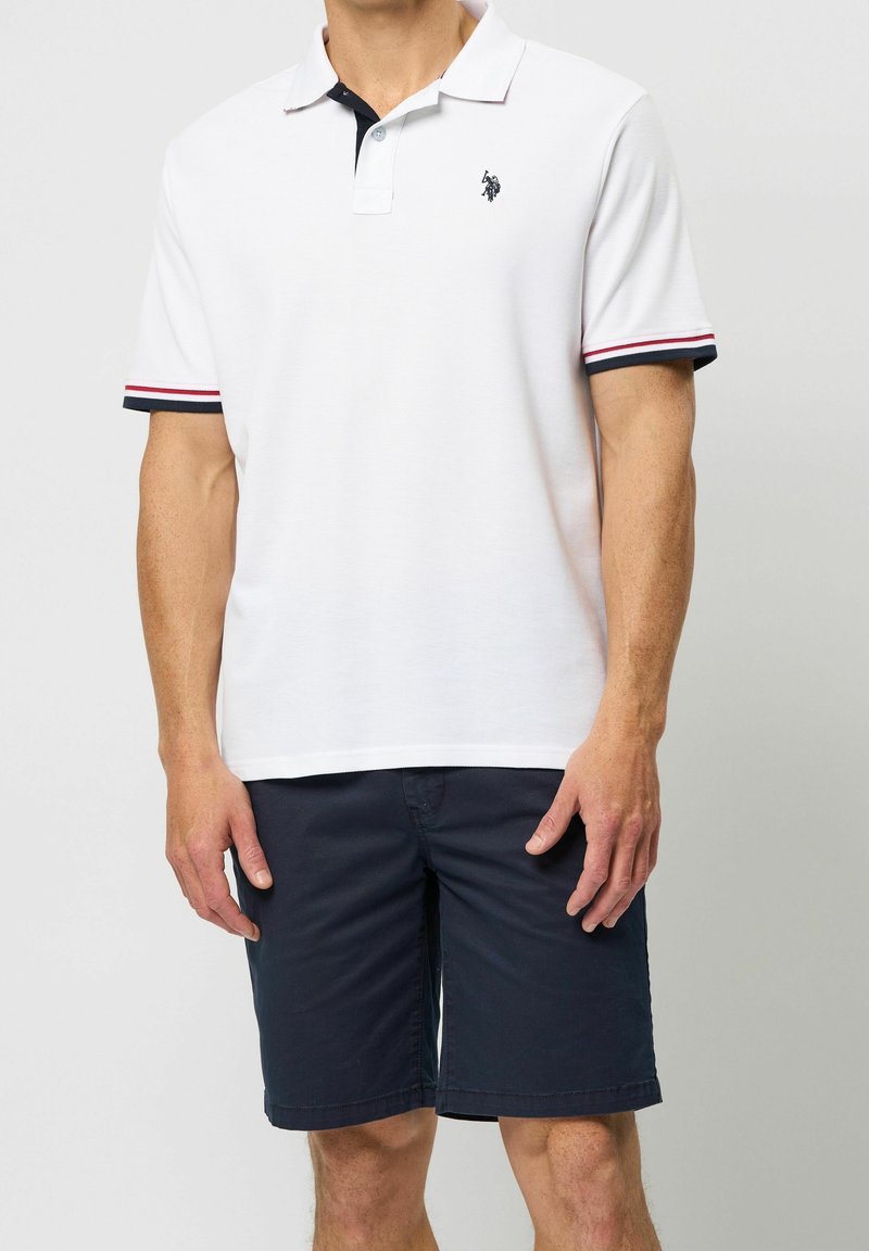 Homme portant un polo blanc à manches courtes avec des bordures rouges et noires et un short bleu marine jusqu'aux genoux, debout devant un fond uni.
