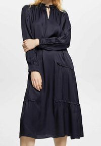 Marinblå midi-klänning i mjukt tyg med långa ärmar, nyckelhålshals och volangdetaljer på fickorna, vilket skapar en mjuk silhuett.