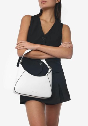 Witte leren handtas met zwarte accenten, gebogen vorm en een enkele schouderriem, vastgehouden tegen een donkere outfit op een neutrale achtergrond.