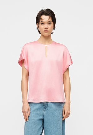 Moterys trumpais tamsiais plaukais, vilkinčios laisvą rožinę trumpomis rankovėmis palaidinę su maža priekyje esančia skyle ir šviesiai mėlynais džinsais, stovinčios prieš balto fono.