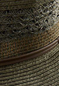 Sombrero de paja verde oliva con un patrón tejido texturizado, que cuenta con una banda de cuero marrón oscuro. Ala ancha con un diseño plano y en capas.