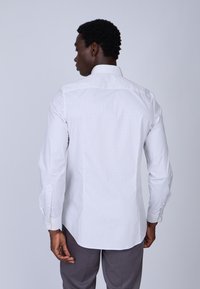 Chemise blanche à manches longues avec de petits pois navy, col standard. Le matériau semble léger et lisse.