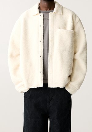 Personne portant une veste polaire crème avec des boutons-pression, une poche poitrine avant, une chemise grise, un pantalon noir et un collier chaîne argenté.