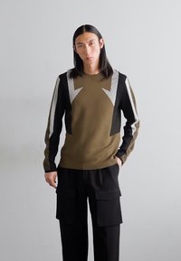 Olívazöld pulóver fekete és szürke geometrikus panelekből, hosszú ujjakkal és kerek nyakkivágással, fekete cargo nadrággal párosítva, mely zsebekkel rendelkezik.