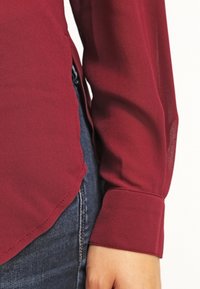 Burgundy blouse with a soft, textured fabric, featuring long sleeves, a curved hem, and a buttoned cuff, paired with dark denim.