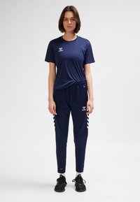 Jonge vrouw staand, gekleed in marineblauwe sportkleding, waaronder een t-shirt en trainingsbroek met witte strepen, en zwarte sneakers op een witte achtergrond.