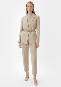 Beżowy garnitur o dopasowanym kroju z marynarką z paskiem i pasującymi spodniami, charakteryzujący się gładką teksturą i ostrymi liniami. Czółenka z lekko szpiczastym noskiem w jasnym kolorze.