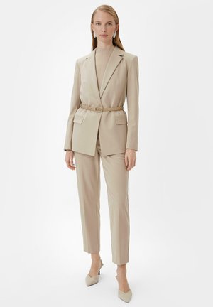 Beżowy garnitur o dopasowanym kroju z marynarką z paskiem i pasującymi spodniami, charakteryzujący się gładką teksturą i ostrymi liniami. Czółenka z lekko szpiczastym noskiem w jasnym kolorze.