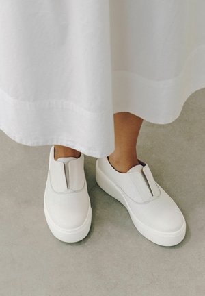 Persona che indossa sneakers bianche slip-on con piattaforma e una gonna bianca, in piedi su un pavimento grigio.