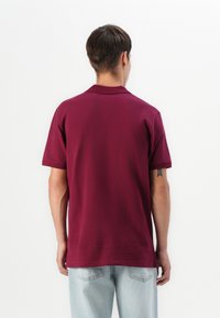 Maronfarvet kortærmet polo t-shirt med klassisk krave, glat tekstur og slidser i siderne. Designet med en afslappet pasform og minimal detaljer.