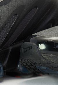 Chaussures de sport noires avec un dessus texturé, des panneaux en maille, un talon arrière brillant et une semelle en caoutchouc à motifs. Logo Nike visible.