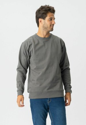 Mand, der kigger til siden iført en grå langærmet rundhalset sweatshirt og blå jeans, stående mod en ensartet lys baggrund.