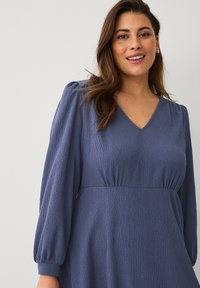 Robe bleue texturée avec un décolleté en V et des manches longues bouffantes, cintrée à la taille, présentant un tissu doux et fluide pour le mouvement.