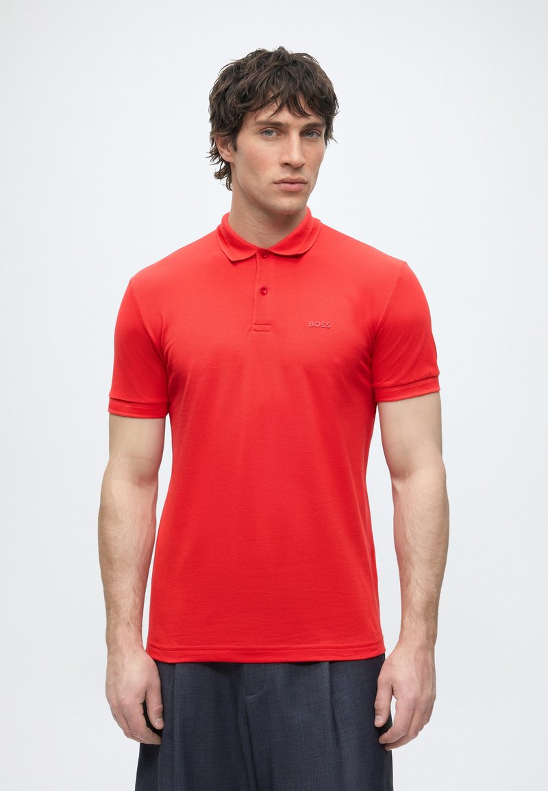 Homme aux cheveux courts et foncés portant un polo rouge à manches courtes et un pantalon foncé, debout devant un fond blanc uni.