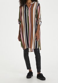 Gestreifte Tunika in verschiedenen Farben mit V-Ausschnitt und Knopfdetails, kombiniert mit schwarzen Slim-Fit-Hosen und schwarzen Sneakers. Mit umgeschlagenen Ärmeln.
