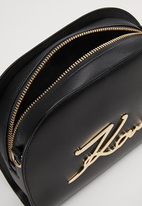 Borsa in pelle nera con due cerniere dorate e un logo dorato prominente sulla parte anteriore. Presenta una forma arrotondata e una superficie liscia.