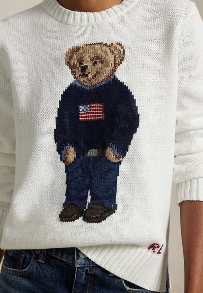 Beli pulover z pletenim medvedkom, ki nosi mornarsko moder pulover z ameriško zastavo in modre kavbojke, nosi oseba v modrih kavbojkah.