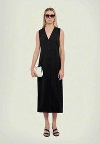 Mulher vestindo um vestido midi preto, sem mangas, com botões, sandálias pretas, óculos de sol e segurando uma pequena bolsa branca contra um fundo simples.