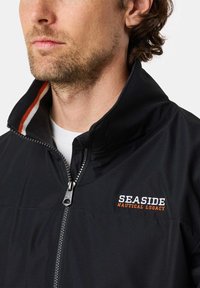 Man draagt een zwart jack met rits, met "SEASIDE NAUTICAL LEGACY" geborduurd op de borst en gestreepte kraagdetail.