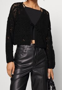 Černý háčkovaný cardigan s otevřeným předním dílem, zavazováním a dlouhými rukávy. Nositý přes černý top a v kombinaci s koženými kalhotami s vysokým pasem.