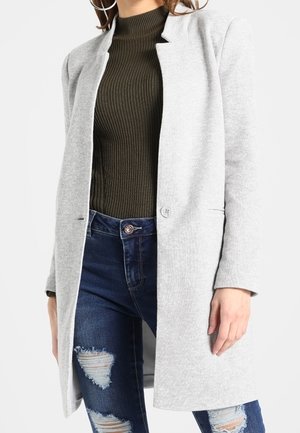 Kvinde iført lys grå lang frakke, mørk ribstrikket rullekrave og revnede blå jeans, stående med venstre hånd afslappet.