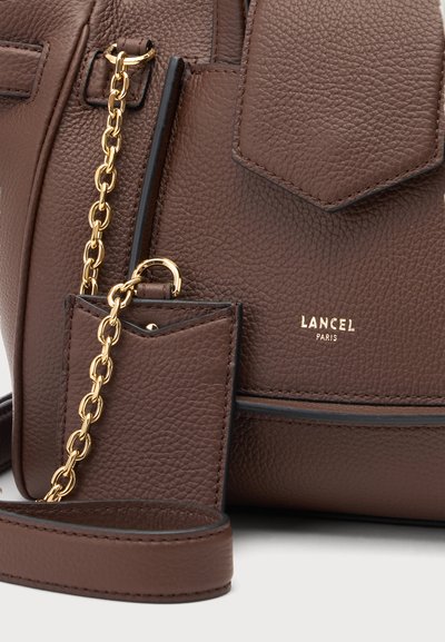 Близък план на кафява чанта Lancel Paris от груба кожа с кафяви зърна, златна верижка, пришит джоб с капаче и малък черен калъф, съвпадащ по стил.