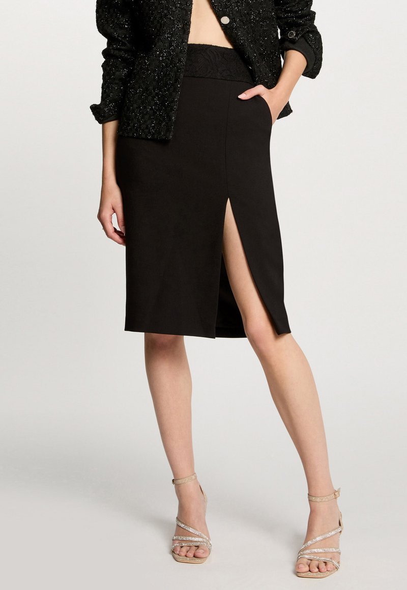 Vrouw in een zwarte rok tot de knie met een voorkeurscheur, gecombineerd met een zwarte textuurjasje en beige sandalen met hoge hakken en bandjes.