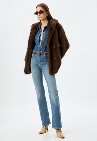 Abrigo de peluche marrón sobre una camisa de mezclilla azul, combinado con jeans acampanados de un azul claro y tacones con estampado de serpiente. Los accesorios incluyen gafas de sol oscuras.