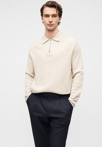 Bež pulover z zadrgo na ovratniku, sproščenega kroja in pleteno teksturo, kombiniran z mornarsko modrimi hlačami z naborki in stranskimi žepi.