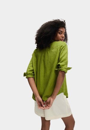 Frau mit lockigen Haaren, die ein hochgekrempeltes langärmliges grünes Hemd und einen weißen Rock trägt, steht mit hinter dem Rücken verschränkten Händen.