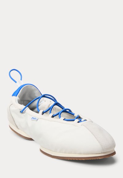 Zapato casual blanco con cordones elásticos azules, lengüeta azul en el talón, puntera suave de ante y suela de goma marrón sobre un fondo claro.