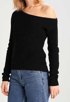 Femme portant un pull noir côtelé à épaules dénudées et un jean bleu sur un fond clair uni.