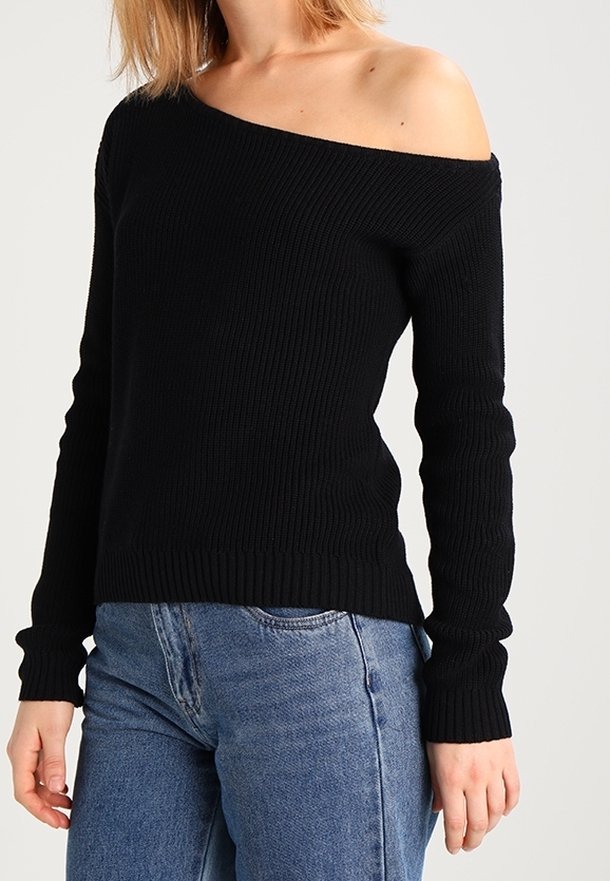 Femme portant un pull noir côtelé à épaules dénudées et un jean bleu sur un fond clair uni.
