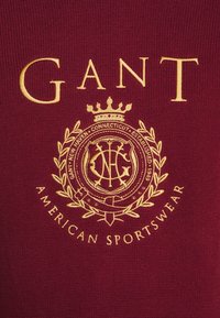 Tissu bordeaux orné d'une broderie dorée avec "GANT", une couronne, et un emblème circulaire portant les inscriptions "NEW HAVEN CONNECTICUT EST. 1949" et "AMERICAN SPORTSWEAR".