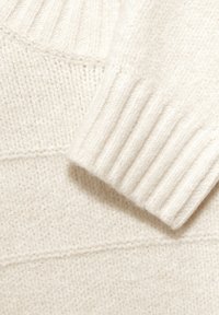 Sezione di un maglione lavorato a maglia color crema con polsini a coste, morbida texture e dettagli di cucitura orizzontale, che mette in mostra un design accogliente.