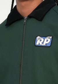 Zelena jakna s zatvaranjem na zip, crnim baršunastim ovratnikom i plavim zakrpom s natpisom "R.P." i crtanih detalja.