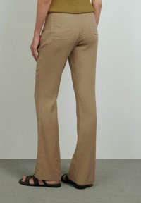 Pantalon évasé beige en tissu doux, avec deux poches arrière et une texture lisse. Associé à des sandales tissées noires.