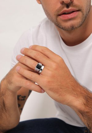 Silver ring featuring a black rectangular stone, smooth finish, and a wide band. Hands resting on each other, skin tone visible.