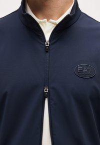 Μπλε ναυτικό μπουφάν με φερμουάρ, δύο φερμουάρ και λογότυπο EA7 Emporio Armani στο στήθος πάνω από λευκό πουκάμισο με γιακά.