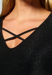 Svart topp med V-ringning, med korsade axelband och en texturerad metallisk finish. Tyget har en subtil glans som förstärker utseendet.