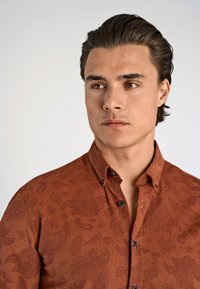Bruine button-up shirt met een textuur van paisleypatroon, voorzien van zwarte knopen en een klassieke kraag. De stof lijkt lichtgewicht en soepel.