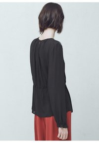 Blusa nera a maniche lunghe con vita arricciata e texture morbida. Presenta una chiusura a chiave sulla schiena e polsini elastici per una vestibilità rilassata.