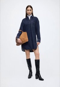 Džínové košeľové šaty tmavomodrej farby, s golierom na gombíky, dlhými rukávmi a jedným náprsným vreckom. Nosené s čiernymi čižmami po kolená a béžovou taškou.