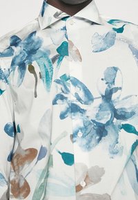 Chemise boutonnée en tissu léger, présentant un motif floral aquarelle dans des tons de bleu et de brun sur fond blanc.