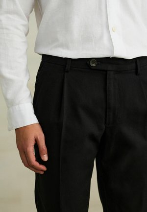 Man wearing black pleated trousers and a white long-sleeve button-up shirt, with left hand resting beside pants.