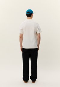 Balta kokvilnas T-krekls ar īsām piedurknēm un redzamām šuvēm; valkāts ar melnām biksēm; modelis valkā zilu cepuri, skatās prom no skatītāja.