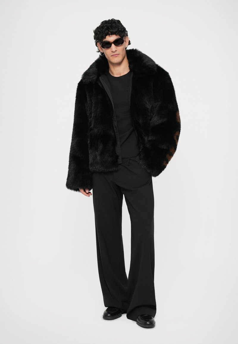 Zwarte nepbontjacket met een hoge kraag, wijde mouwen, gecombineerd met een zwart top en een wijduitlopende broek. Model draagt zwarte zonnebril.