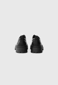 Paire de chaussures noires en cuir avec semelles épaisses à gros crampons, vues de dos, avec languettes de traction et semelles en caoutchouc épaisses à relief, sur fond gris.
